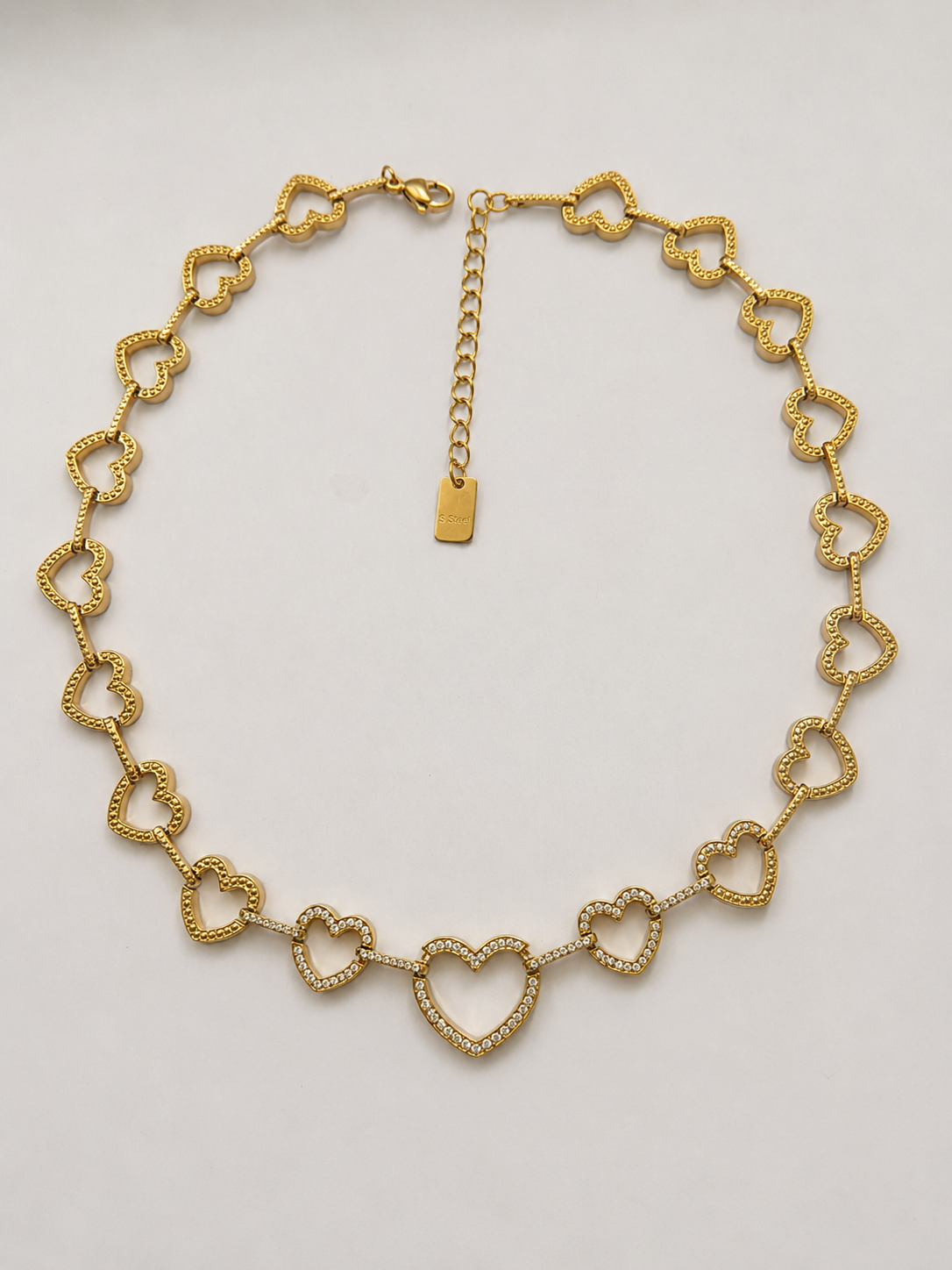 Collier Athéna en packshot, cœurs ajourés dorés, cœurs du centre pavés de zircons, fermoir mousqueton, plaquette S.Steel